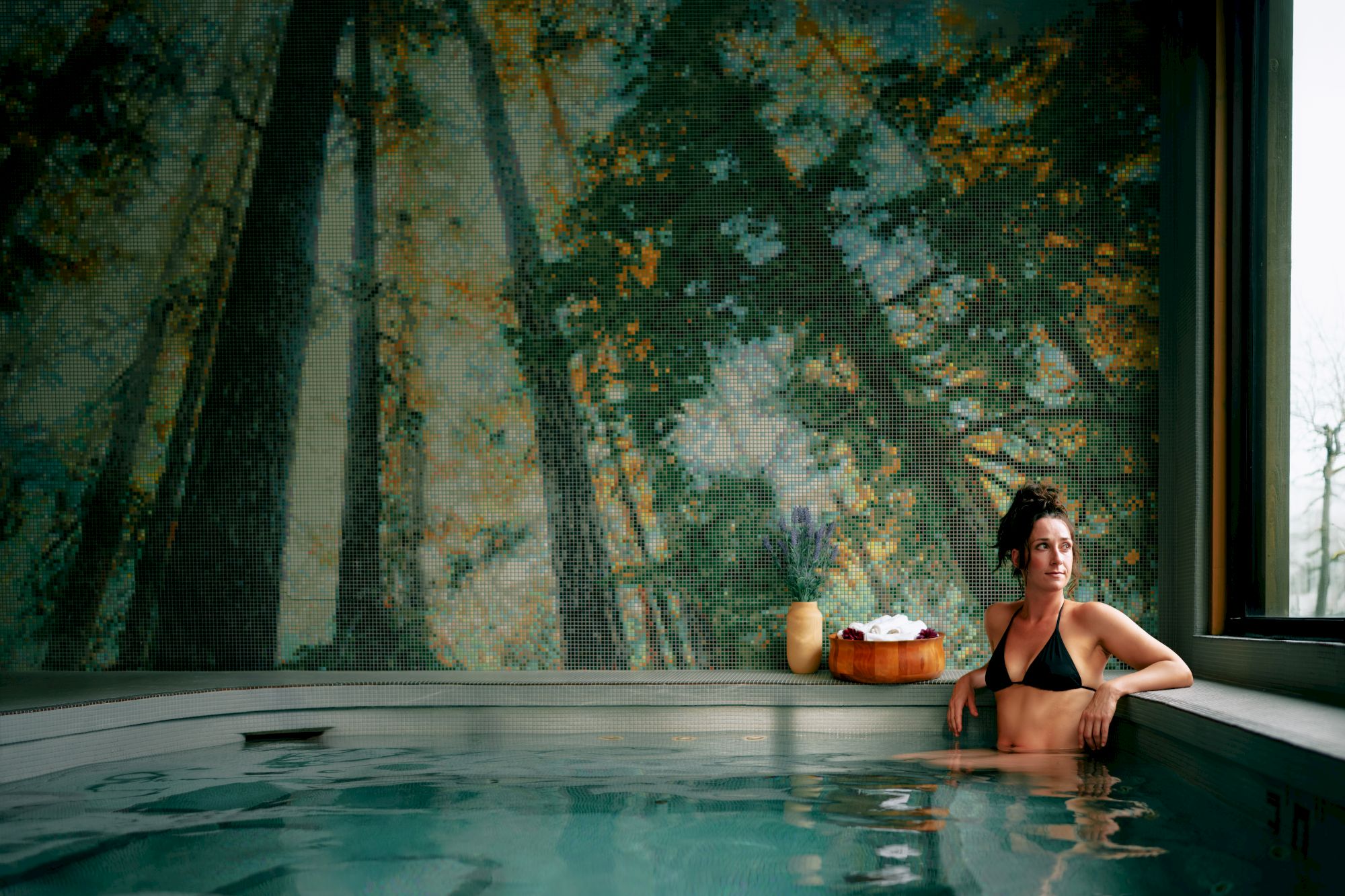 Person lounging in an indoor pool, surrounded by colorful mosaic tiles and a flower vase on the ledge.