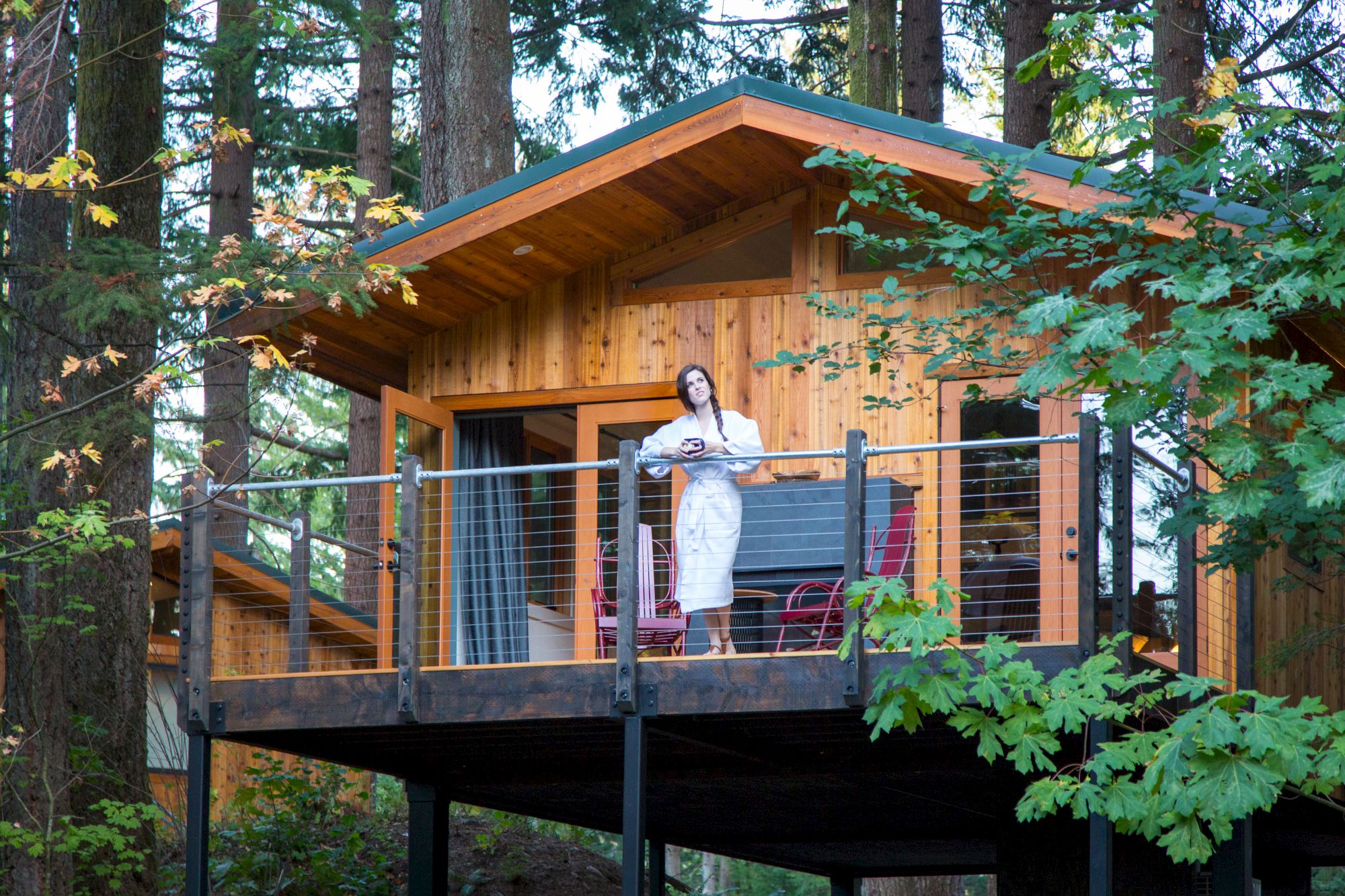 A woman in a white dress stands on a wooden deck in a forest, framed by trees and a metal railing.