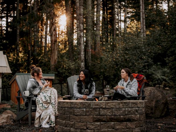 Three women sit around an outdoor fire pit, laughing and toasting with wine in a forest setting at sunset.