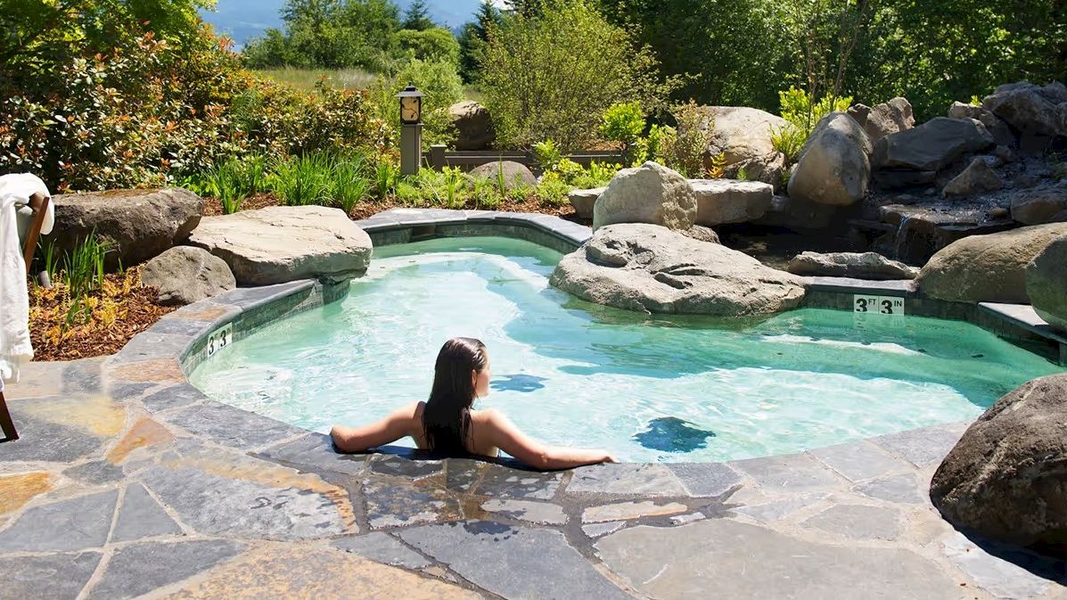 A person relaxes in a rock-lined outdoor pool surrounded by lush greenery under a clear sky.
