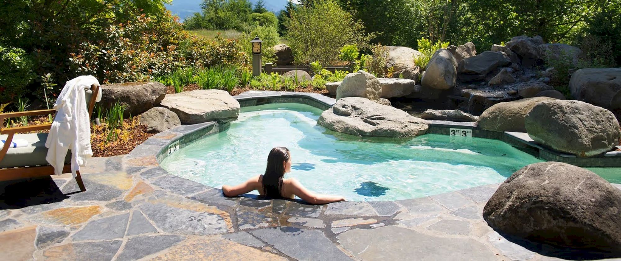 A person relaxes in a rock-lined outdoor pool surrounded by lush greenery under a clear sky.