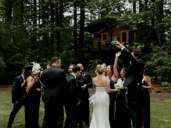 A bride in a white gown and her wedding party cheer in a lush forest garden, posing on the lawn near a cabin.