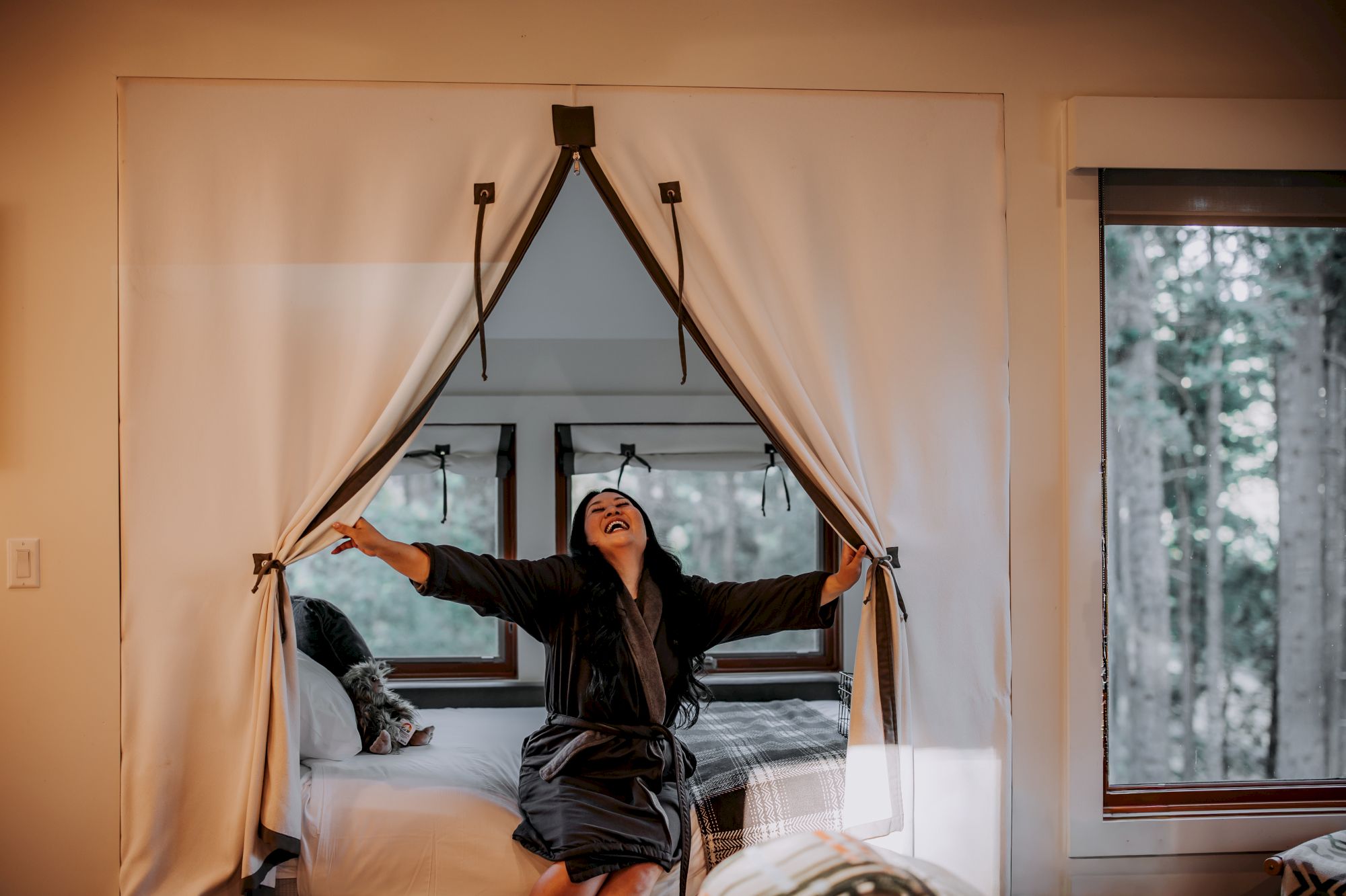A woman in a robe playfully holds open airy curtains above a bed, with a dog nearby in a bright, cozy room.