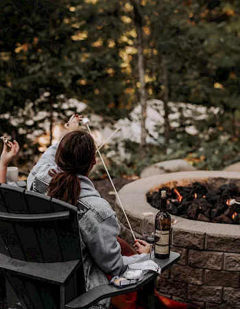 Three people sit by a backyard fire, roasting marshmallows on skewers and chatting at dusk.