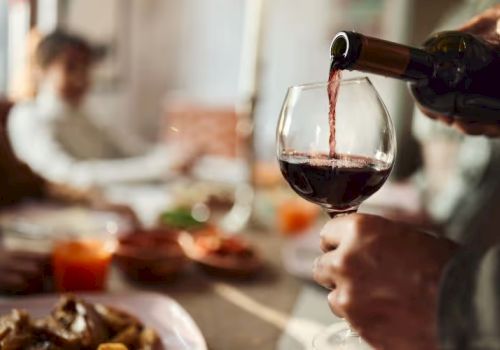A person is pouring red wine into a glass at a dining table, with blurred figures and food in the background, creating a cozy atmosphere.