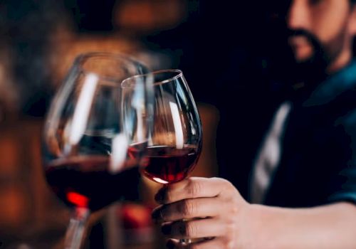 Several glasses of red wine are being clinked together in a toast, held by a person in a blurred background setting.