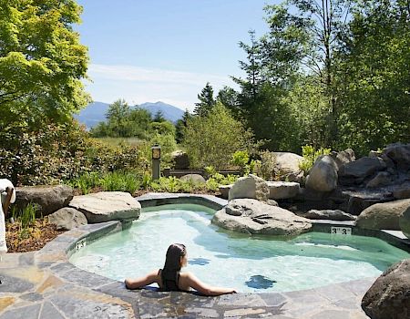 A person relaxing in an outdoor hot spring pool surrounded by rocks, lush trees, and distant mountains under a blue sky.