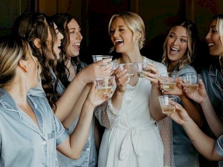 A group of women in pajamas laugh and toast together, clinking plastic cups with drinks at a party.