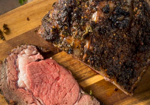 A sliced, peppery herb-crusted roast beef sits on a wooden board, with dark seasoned crust and juices visible.