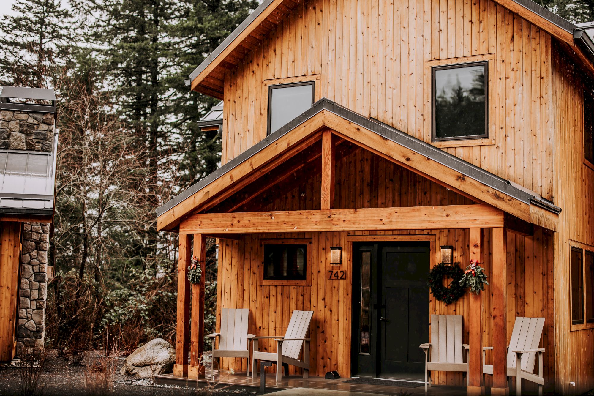 A modern wooden cabin exterior with a stone chimney, large windows, black door, porch chairs, and wreaths in a snowy forest setting.