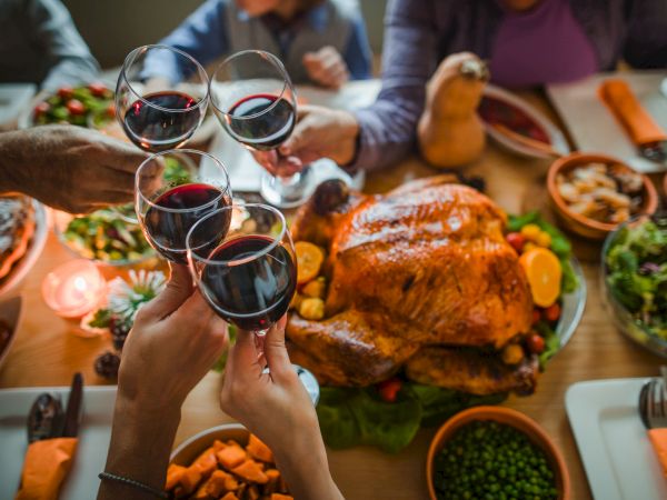 A group cheers with red wine glasses over a festive dinner table piled with roasted meat and colorful vegetables.