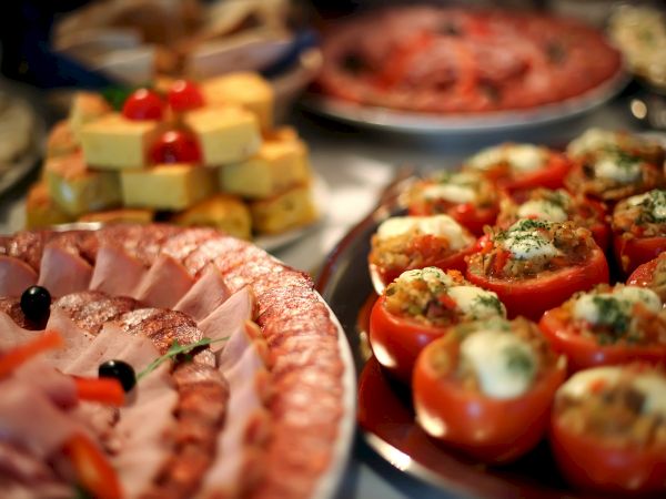 A buffet spread of sliced cured meats, stuffed tomatoes, cheeses, and veggies with herbs on top.