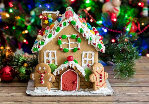 A decorated gingerbread house with gumdrop shingles and icing details sits on a holiday table, surrounded by colorful Christmas lights.