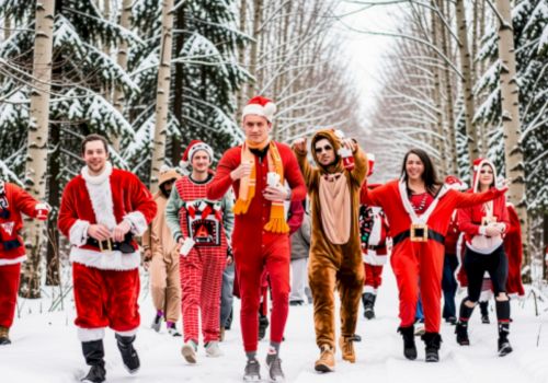 A group of people in Santa, elf, and reindeer costumes walk through a snowy forest, smiling and waving.