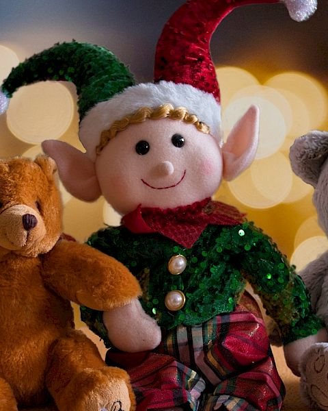 A smiling elf in a green sequined suit sits with three plush teddy bears&mdash;brown, gray, and cozy at a holiday table.