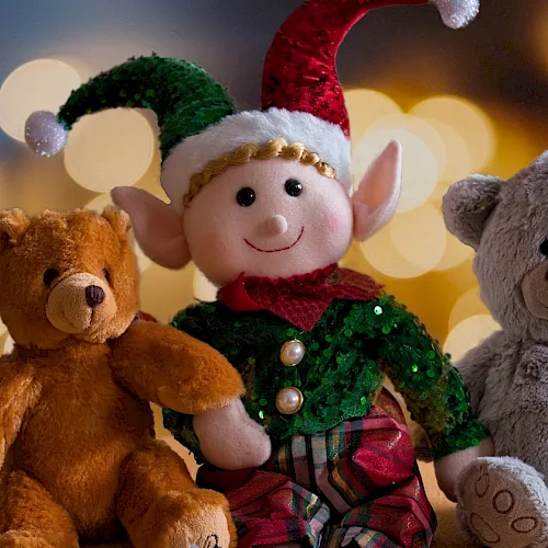 A smiling elf in a green sequined suit sits with three plush teddy bears—brown, gray, and cozy at a holiday table.