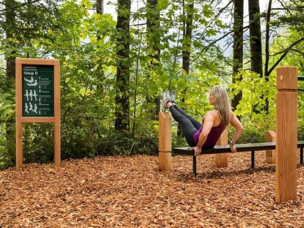 A person exercises on outdoor fitness equipment surrounded by trees, with a sign showing workout instructions in a park setting.