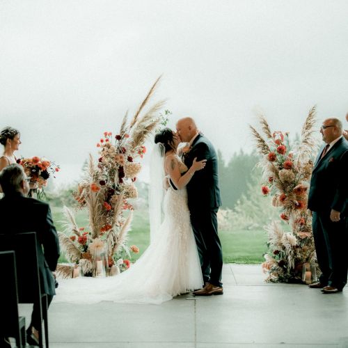 A couple kisses at a wedding ceremony, surrounded by bridesmaids and groomsmen, with floral decorations and guests watching.