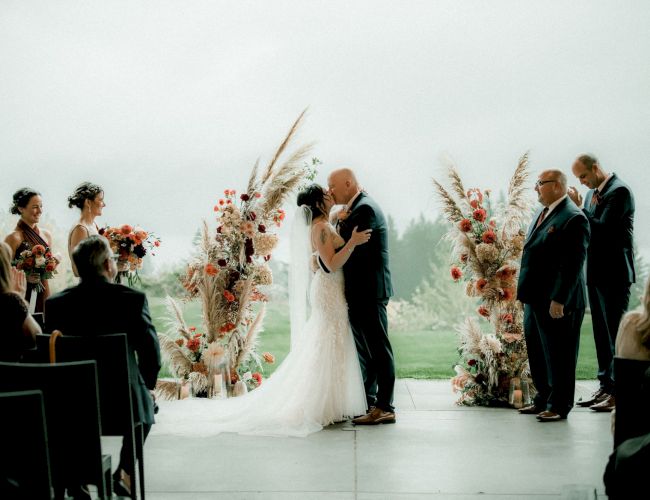 A couple kisses at a wedding ceremony, surrounded by bridesmaids and groomsmen, with floral decorations and guests watching.