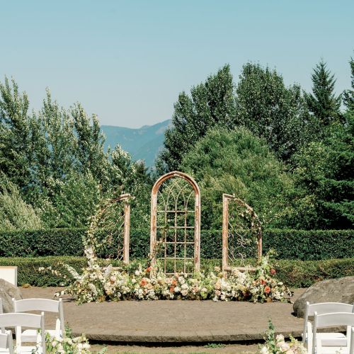 An outdoor wedding setup with a rustic arch, floral arrangements, and white chairs, set against a backdrop of lush greenery and mountains.