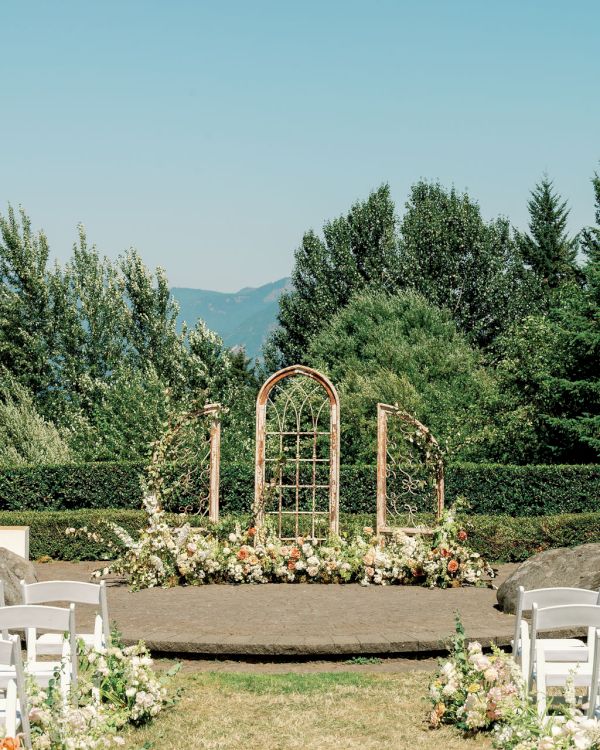 An outdoor wedding setup with a rustic arch, floral arrangements, and white chairs, set against a backdrop of lush greenery and mountains.