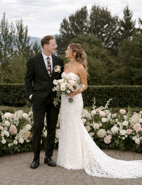 A couple stands together in wedding attire, surrounded by lush greenery and white floral arrangements.