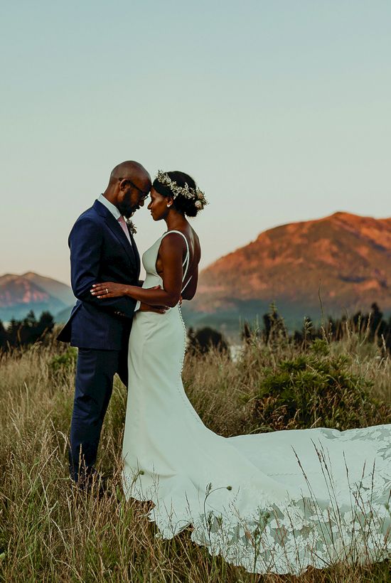A couple in wedding attire embraces in a meadow with mountains in the background during sunset.