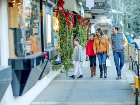 A family strolls through a festive street, shopping by a shop window decorated with lit greenery and red bows.