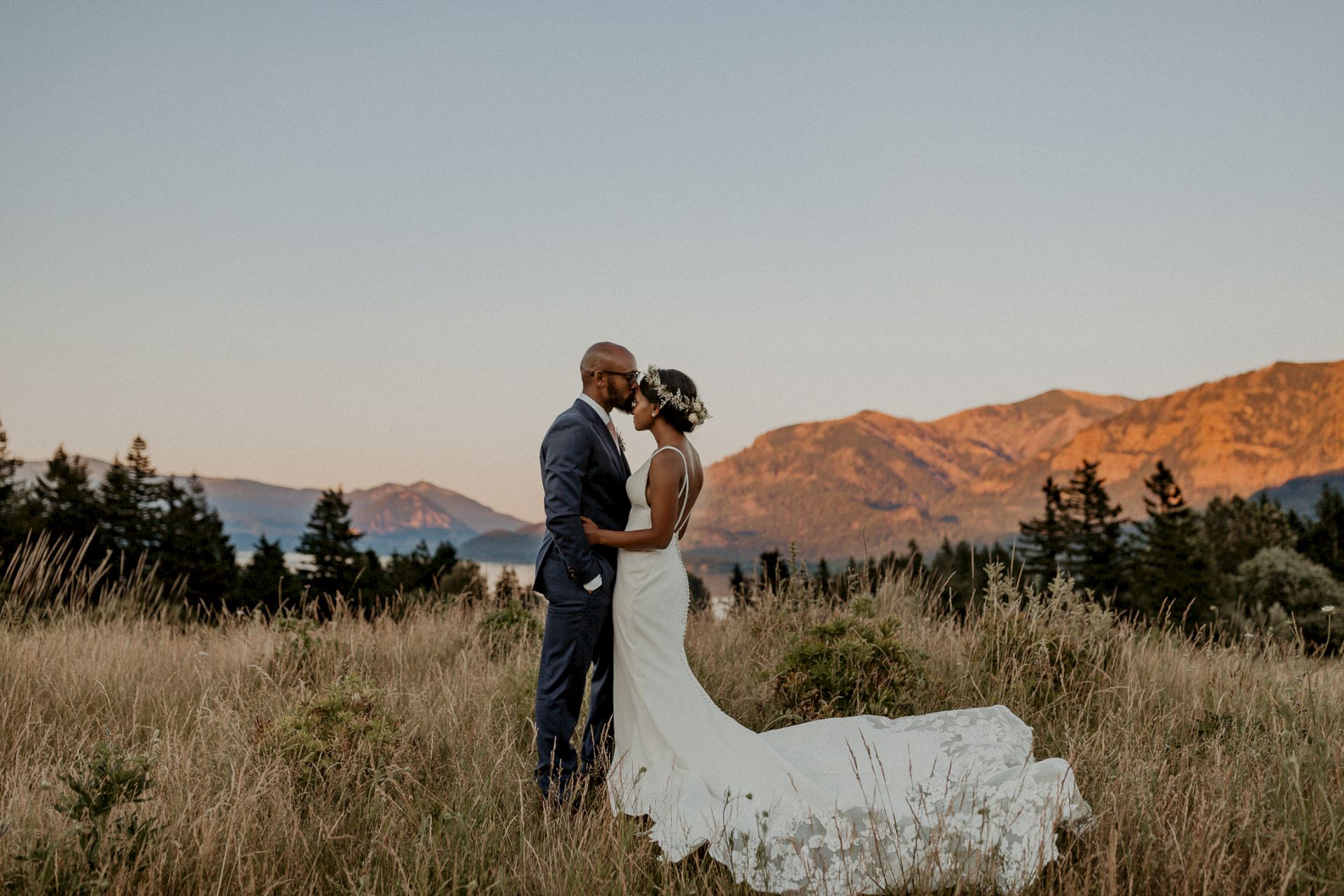 A bride and groom share a kiss outdoors in wedding attire, standing in a grassy field with mountains glowing at sunset.