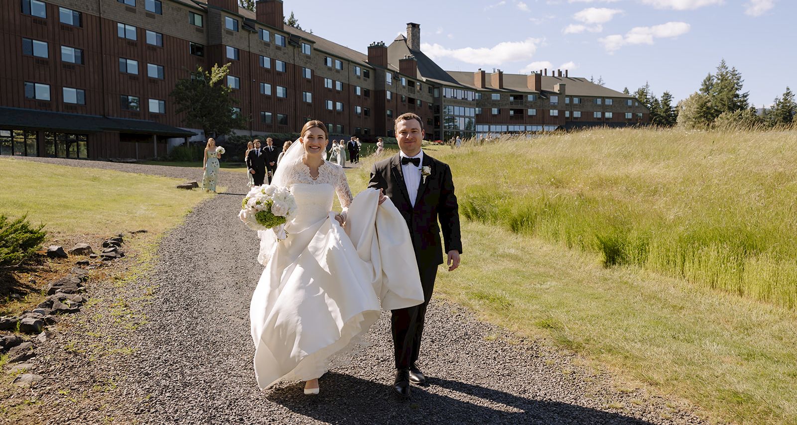A bride and groom in formal wear walk along a gravel path outside a lodge on a sunny day.