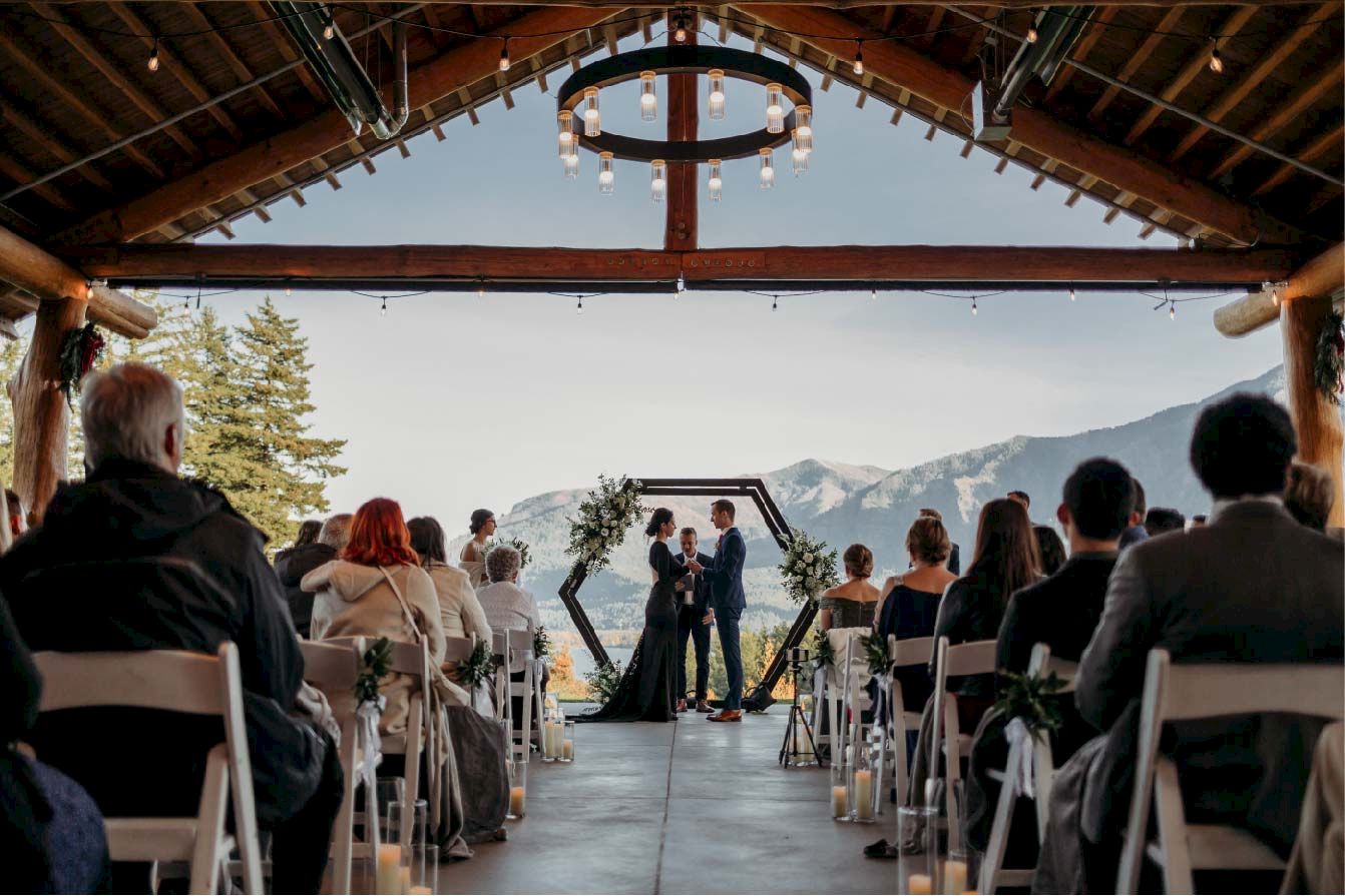 An outdoor wedding ceremony on a terrace with guests seated, a couple at the aisle, and mountains in the background.
