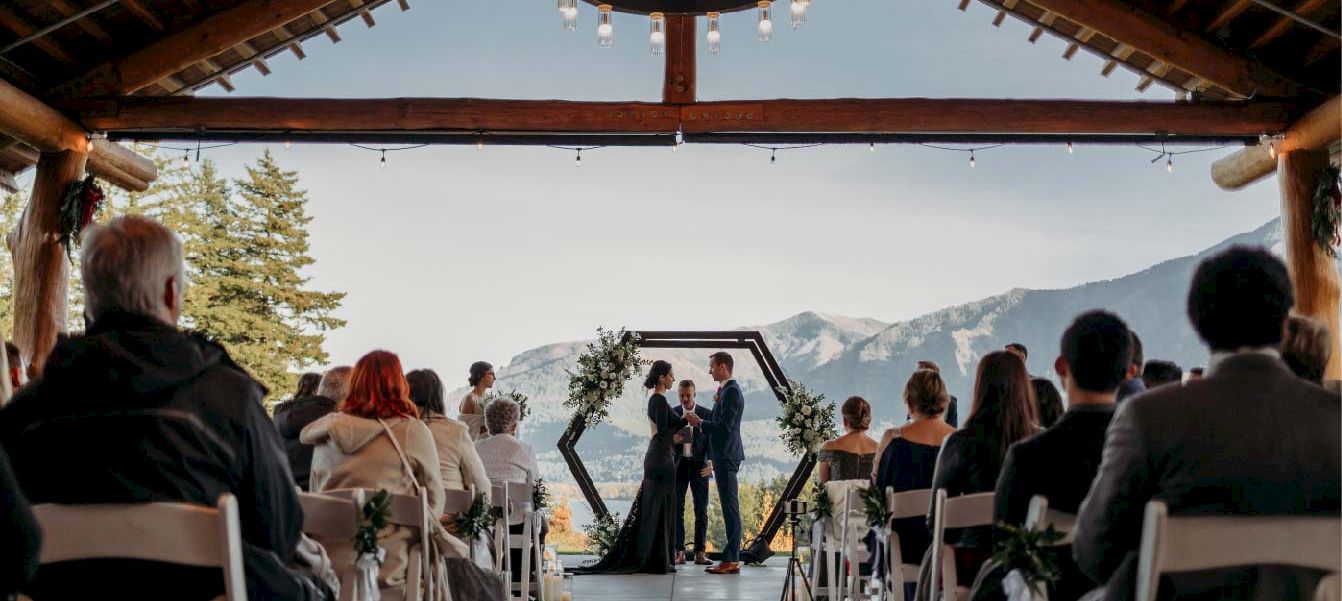 An outdoor wedding ceremony on a terrace with guests seated, a couple at the aisle, and mountains in the background.