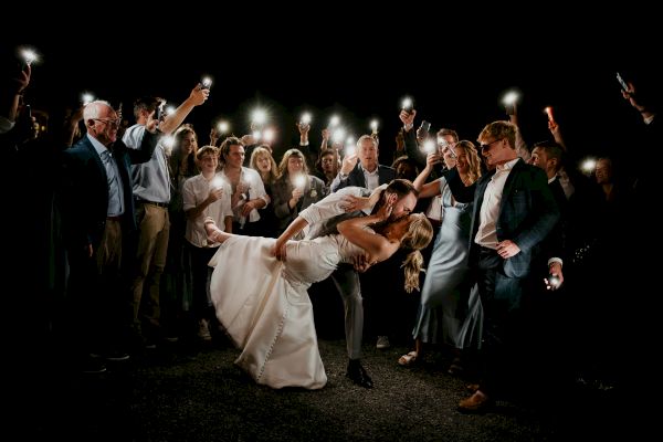 A couple kisses in the center while surrounded by a group holding up lights, creating a celebratory atmosphere.