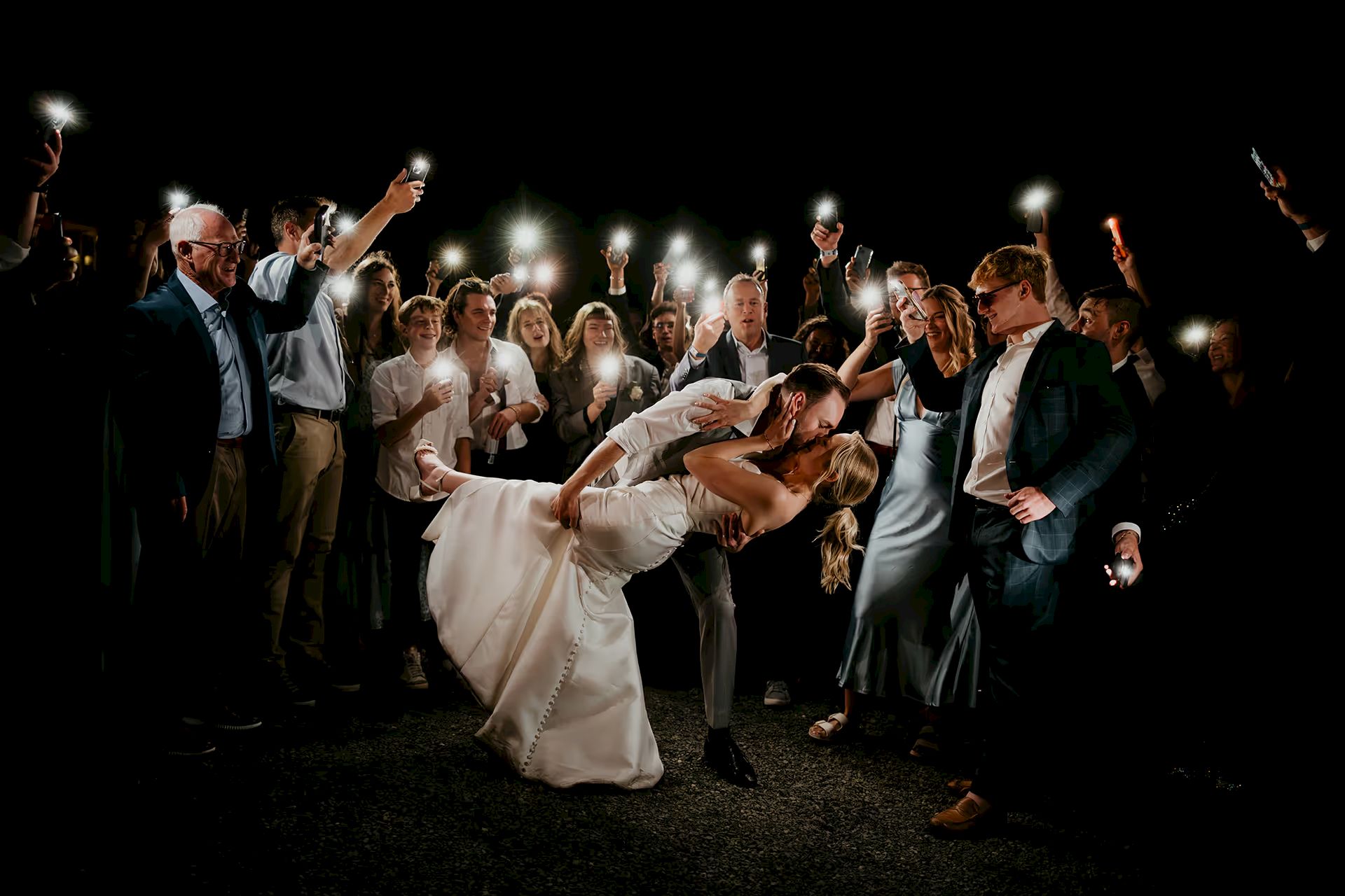A couple kisses surrounded by a crowd holding up phone lights, creating a romantic nighttime scene.