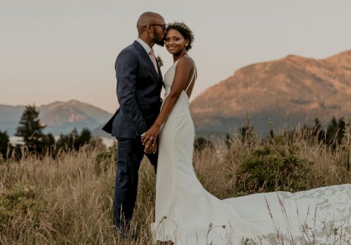 A bride and groom pose outdoors in a grassy field with mountains in the background, smiling and holding hands together at sunset.