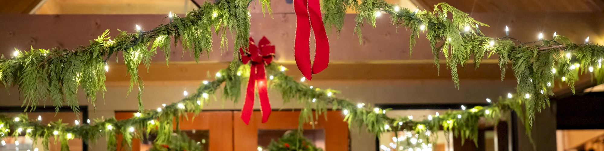 A Christmas garland with tiny lights and a big red bow hangs above doors indoors.