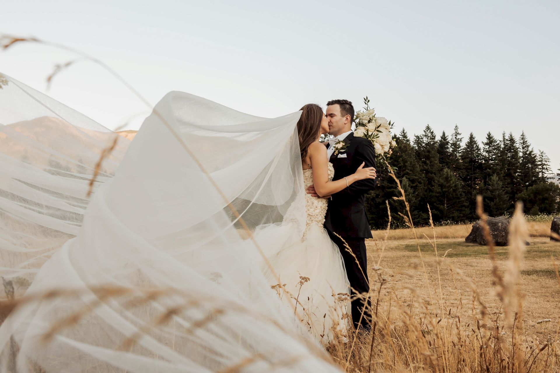 A bride and groom kiss in a field at sunset, their white veil billowing, as they hold a bouquet of cream flowers.