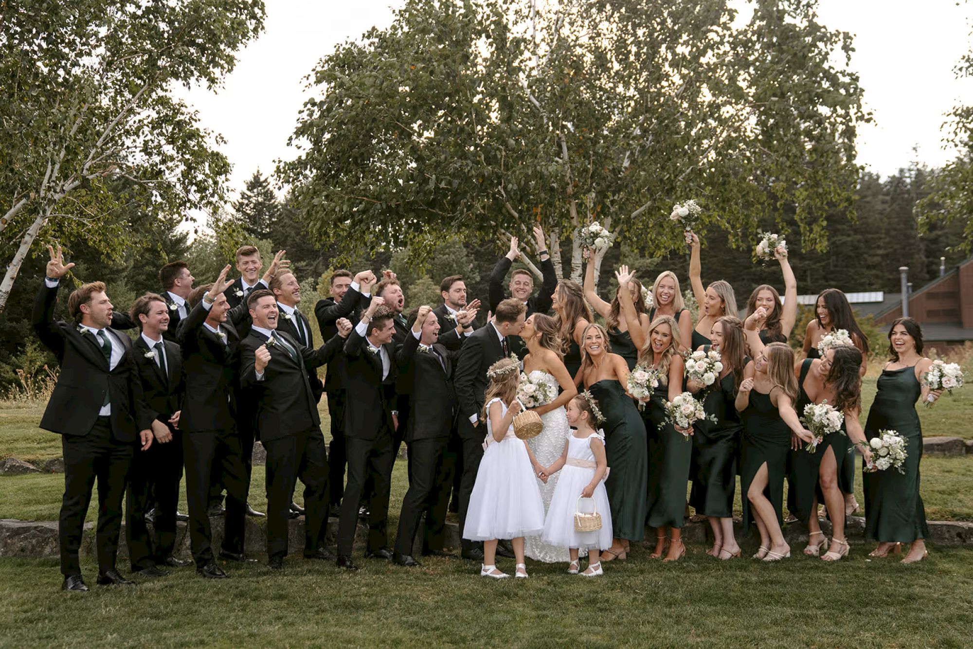 A large wedding party poses outdoors, with bridesmaids in green dresses and groomsmen in suits, celebrating and holding bouquets.
