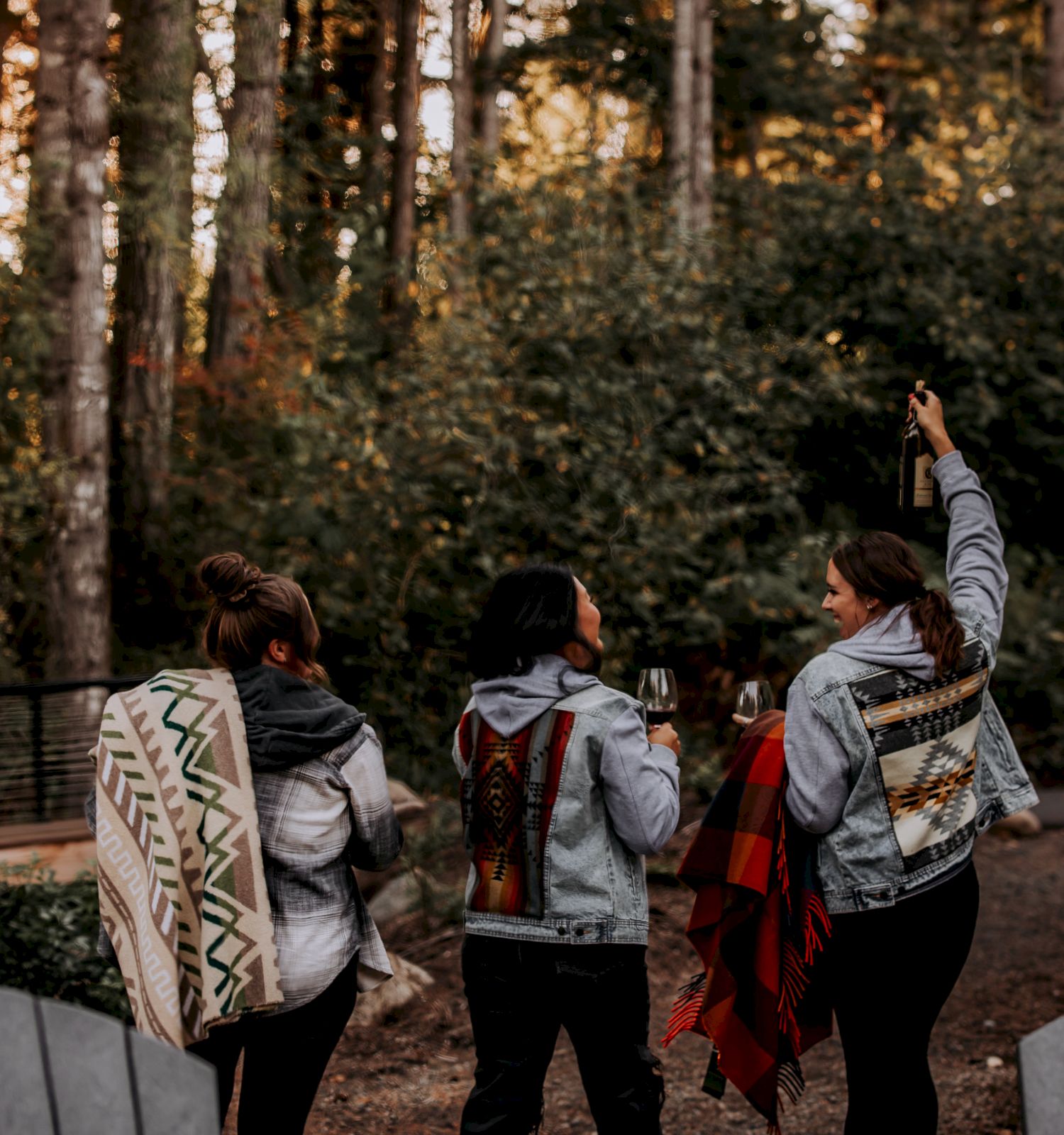 Friends in a forest, toasting with wine while raising a bottle at sunset, wrapped in patterned shawls.