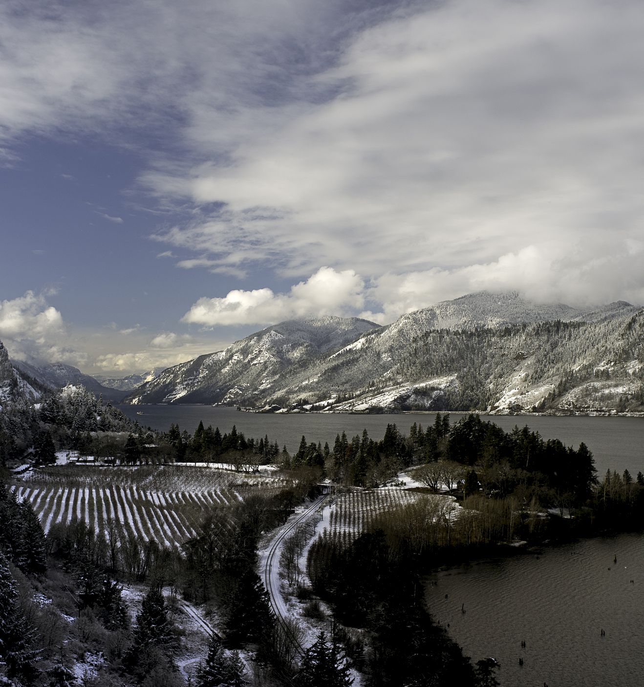A snowy mountain valley with frosted pine trees, a winding road, and a frozen lake under cloudy winter skies.