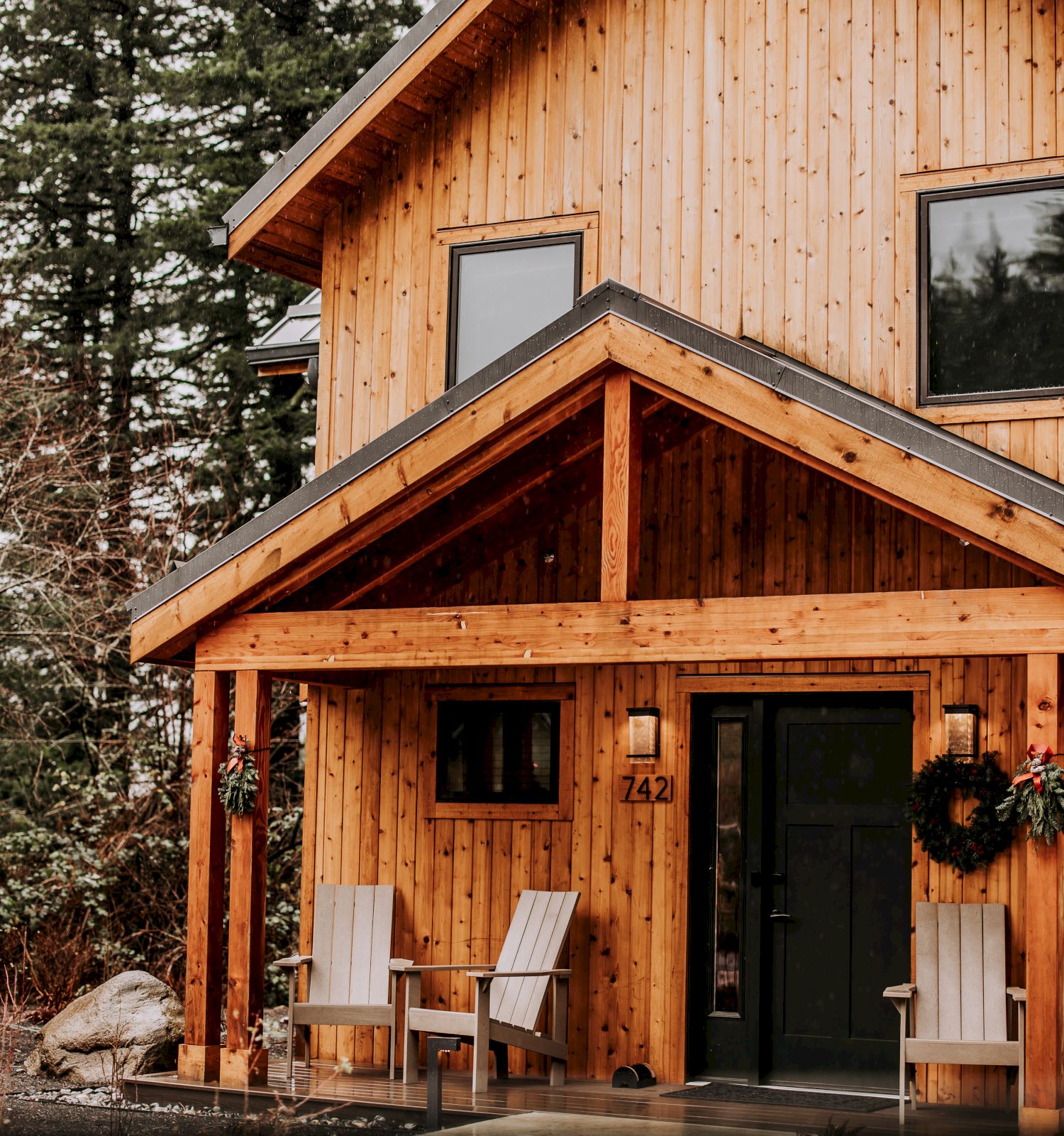 A modern wooden cabin exterior with black doors, large windows, wreaths, and stacked chairs on a porch.