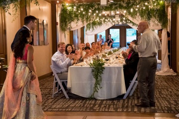 A formal gathering with people seated at a long table, one person standing and speaking, under string lights and greenery decorations.