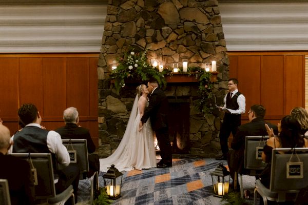 A couple is kissing at a wedding ceremony in front of a stone fireplace, with an officiant and guests present in an indoor setting.