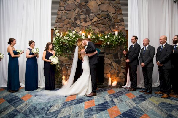 A bride and groom kiss at a wedding ceremony, surrounded by the bridal party. The backdrop is a stone wall with floral arrangements and candles.
