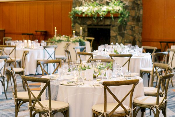 A decorated event space with round tables, white tablecloths, wooden chairs, and a stone fireplace adorned with greenery.