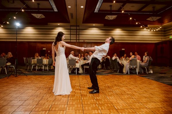 A couple dances energetically on a wooden floor in a decorated hall with seated guests watching and lights hanging above.