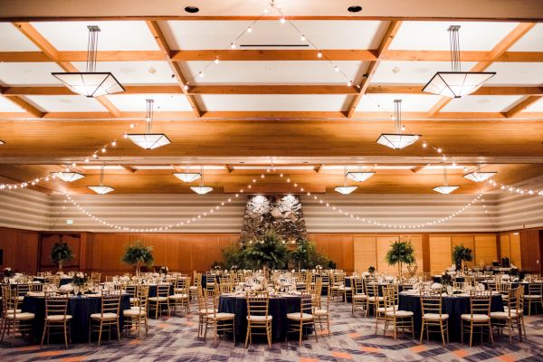 A decorated banquet hall with round tables, chairs, string lights, and a stone wall centerpiece, likely prepared for an event or reception.