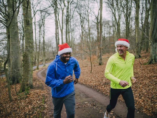 Two runners in winter jackets jog along a forest path, smiling and wearing red-and-white Santa hats in the chilly woods.