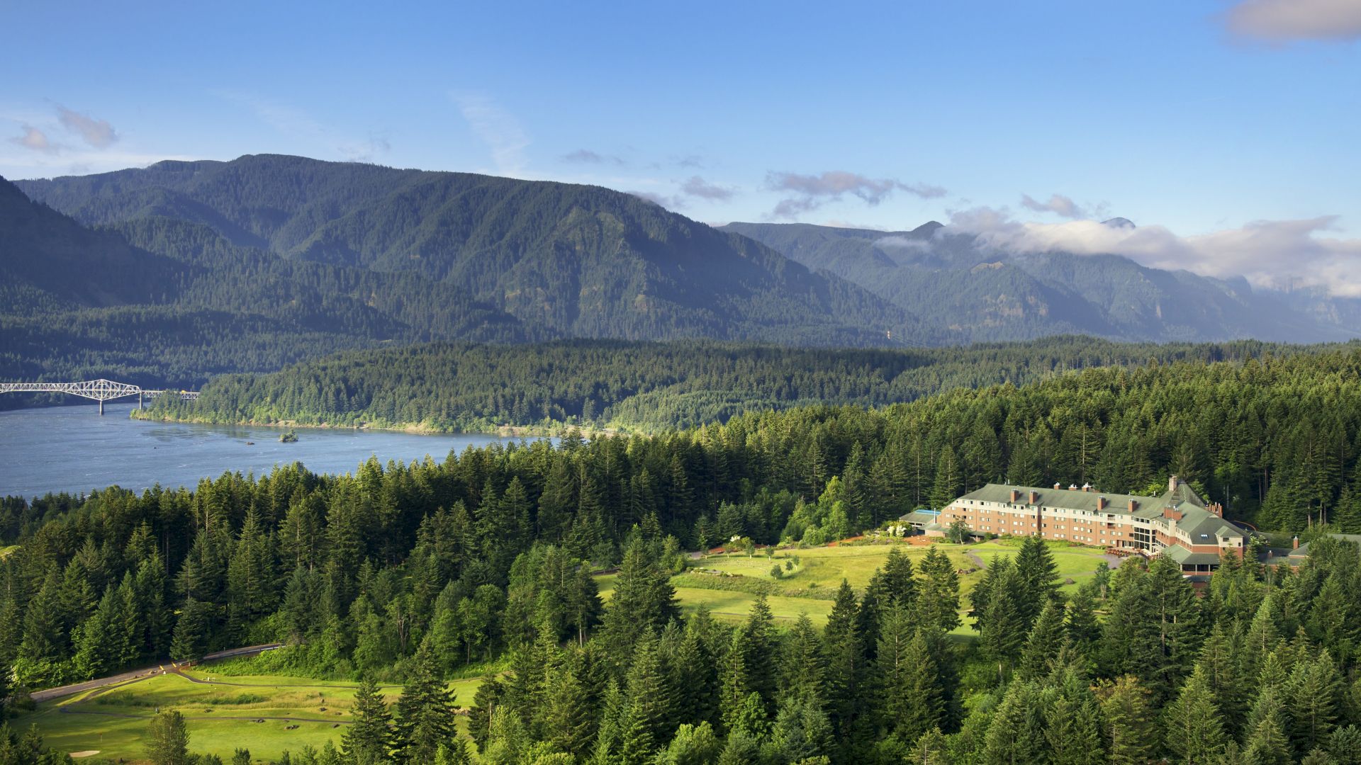 A scenic mountain valley with a river, a truss bridge, dense pine forests, and a hillside resort building beneath misty peaks.