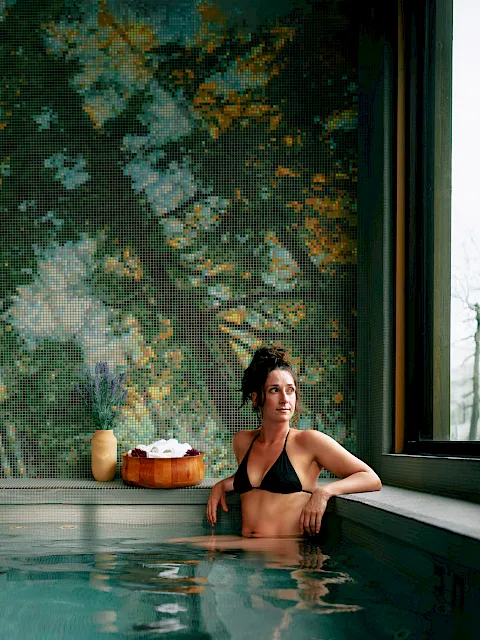 A woman relaxes in an indoor spa pool beneath a colorful mosaic wall, with towels and flowers on the ledge nearby.